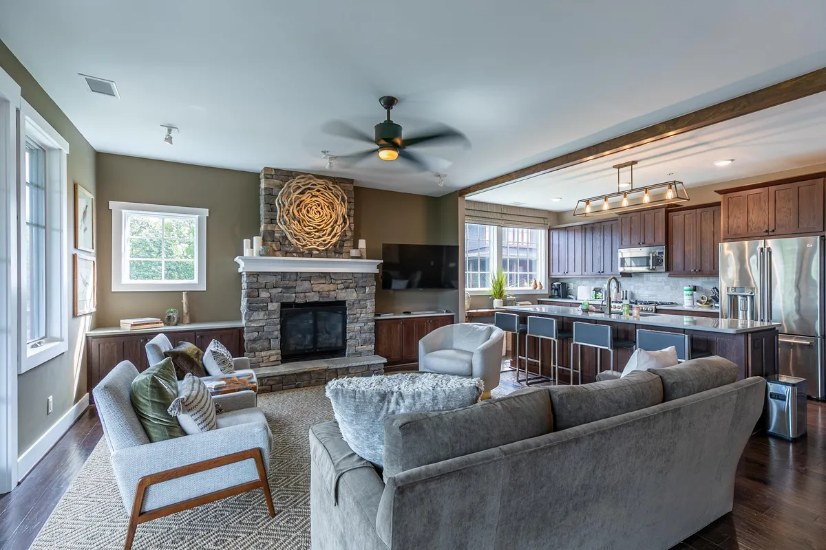 This image captures a spacious open-concept living and dining area featuring a stunning stone fireplace, large windows that flood the room with natural light and offer mountain views, a kitchen island, and tasteful decorative wall art. The overall mood is cozy and welcoming, with comfortable furnishings and thoughtful layout.