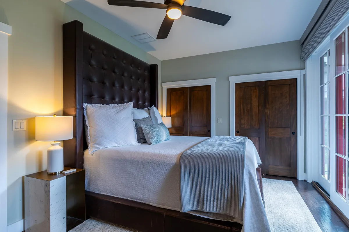 A tranquil bedroom featuring a large tufted headboard, plush bedding, a lamp on the nightstand, and wooden doors. The ambiance is serene and restful, with soft lighting that invites relaxation and comfort.