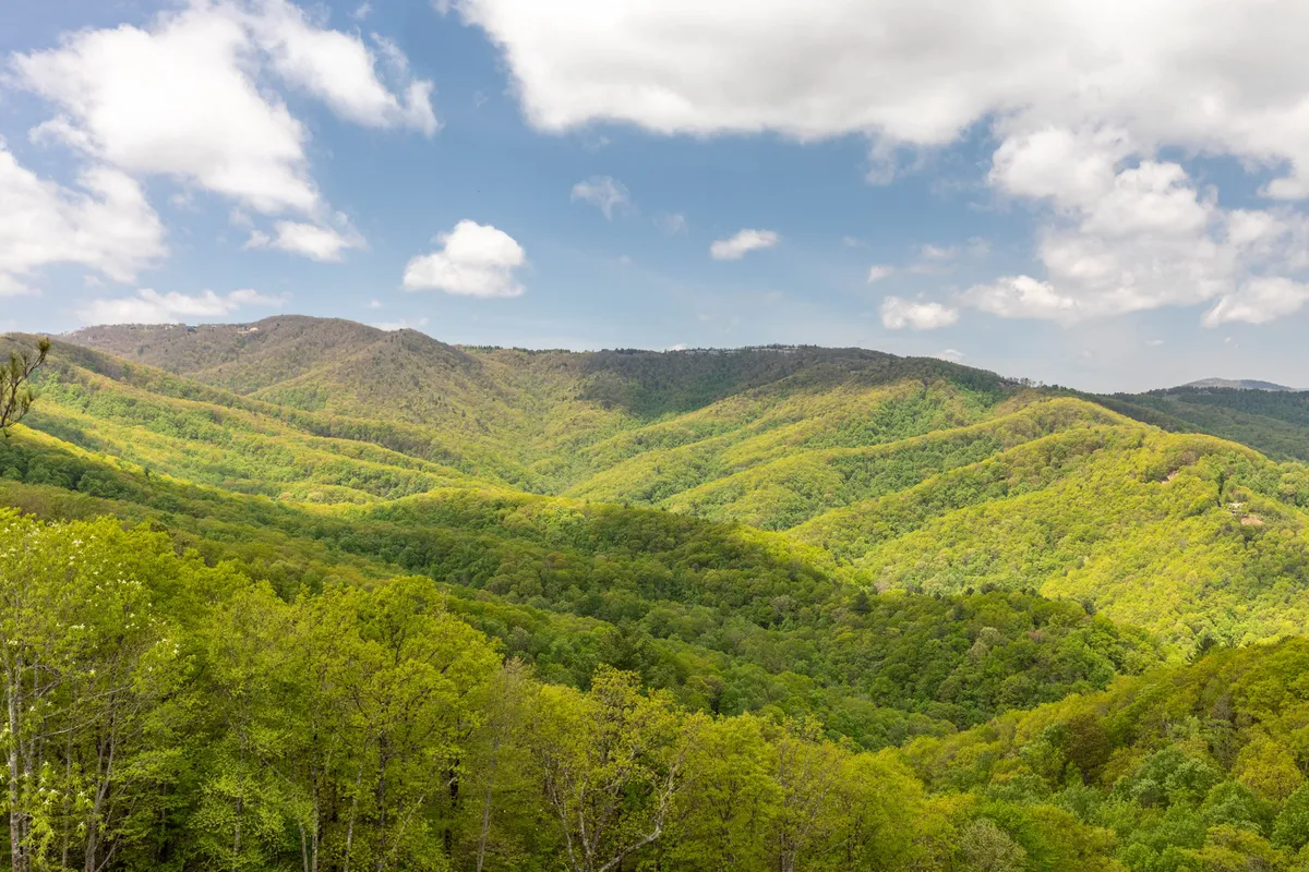 A wide landscape of rolling green hills, distant mountains, and a blue sky with fluffy clouds. The scene invites exploration, hiking, and taking energizing breaks in nature.