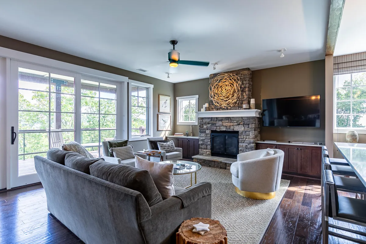 A comfortable living area with a plush sofa, armchair, a coffee table, a stylish fireplace, and expansive windows letting in abundant natural light. The view through the windows opens to the scenic outdoors, making the space feel airy and connected to the landscape.