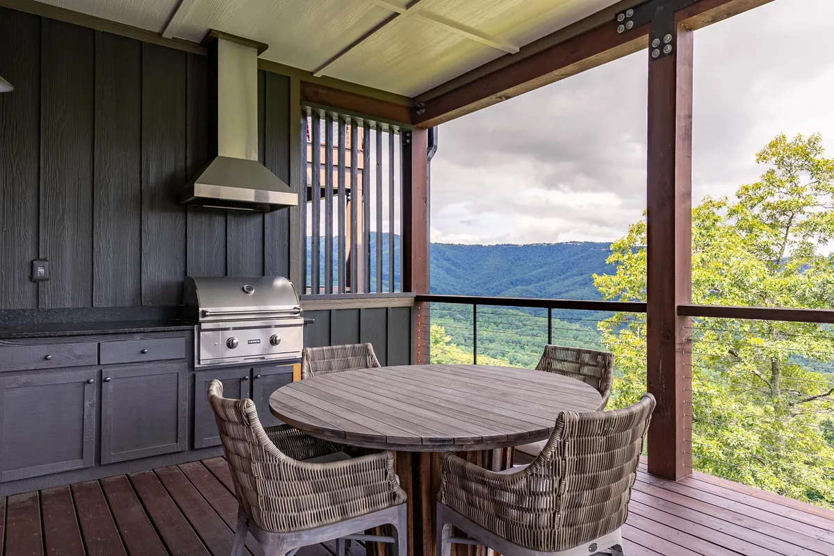 An outdoor dining deck featuring a dining table set for a meal, a built-in grill, and panoramic mountain views. Comfortable seating invites guests to enjoy meals while taking in sunsets over the mountains.