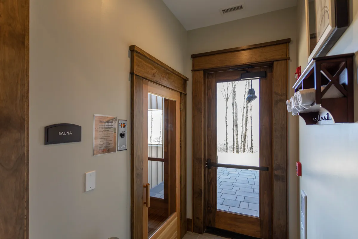 This tranquil interior scene highlights a sauna with its wooden door slightly ajar, offering a glimpse of the serene exterior beyond and soft indoor lighting that creates a calming atmosphere. The space invites relaxation and rejuvenation after a workout.